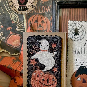 May include: A rectangular punch needle textile art piece featuring a white ghost and orange pumpkins on a black background, mounted on a book. The text reads Friendly Ghost, punchneedle by Teresa Kogut, PN372, and Ann Page on a tin below.