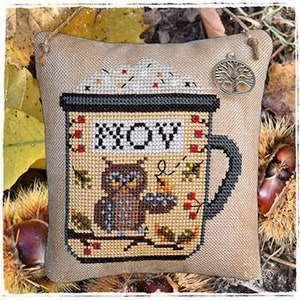 May include: A brown owl with yellow eyes sits on a branch inside a brown and white mug with the word "HOOV" written on it. The mug is filled with a white substance and red berries. The mug is on a beige fabric background.
