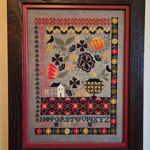 May include: Framed cross-stitch artwork featuring a floral and geometric design in red, yellow, black, and gray. The artwork includes a house, birds, and the alphabet. The frame is dark brown with a red inner border.