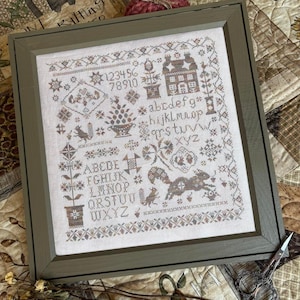 May include: A framed cross-stitch sampler on off-white fabric featuring brown and muted-tone thread designs. The embroidery includes the alphabet in uppercase and lowercase letters, numbers 1-0, a house, squirrels, floral motifs, and geometric borders.