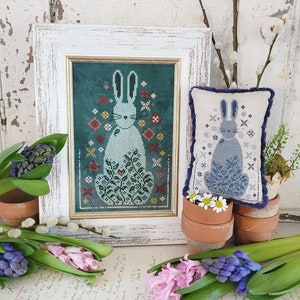 May include: Two framed cross-stitch embroidery designs featuring a blue bunny surrounded by flowers. The bunny on the left is surrounded by a variety of colorful flowers on a teal background. The bunny on the right is surrounded by white flowers on a white background. Both designs are framed in white wood frames.