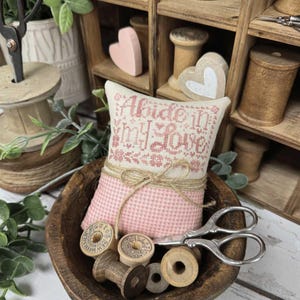 May include: A pink and cream-colored pincushion with the text "Abide In My Love" sits in a wooden bowl with spools of thread and scissors. The pincushion is tied with twine. The words "Primrose Cottage" are at the bottom.