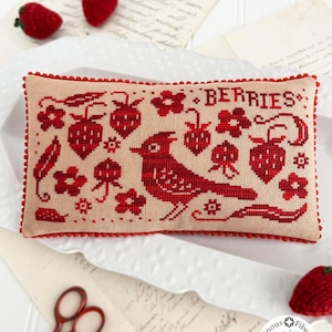 May include: A rectangular, cross-stitched pillow with a red bird and the word "BERRIES" embroidered on a beige background. The design includes red strawberries and flowers. The pillow is trimmed with red pom-poms. Crochet strawberries and vintage paper are in the background.