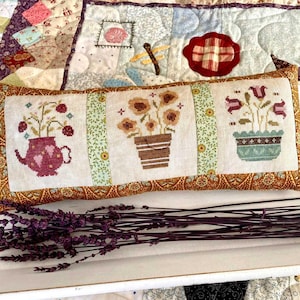 May include: A decorative pillow with a white background and three embroidered images of potted plants. The pillow has a brown and gold border. The pillow is sitting on a white surface with a bunch of dried lavender.