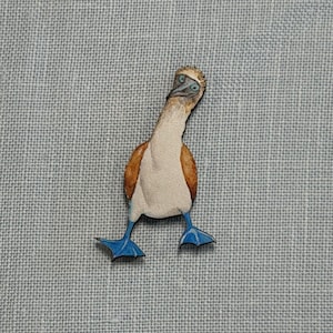 May include: A wooden pin depicting a blue-footed booby bird. The bird has a white body, brown wings, and bright blue feet. The pin is set against a light blue textured background.