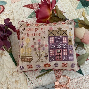 May include: A sampler house cross stitch pattern on a cream pillow with pink and purple accents. The pillow is on a white quilt with a floral pattern and a pair of pink scissors.
