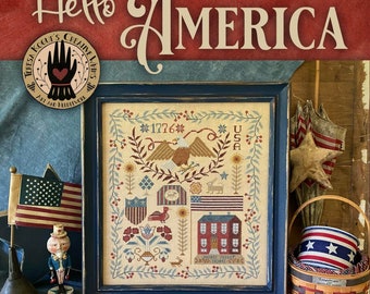 Pode incluir: Um design de ponto cruz emoldurado com temas patrióticos, incluindo uma águia, bandeiras e o texto "1776" e "USA". O design está em uma moldura azul, com as palavras "Hello America" acima. Itens adicionais incluem uma pequena bandeira e uma cesta decorativa.