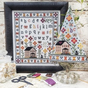 May include: A cross-stitch sampler with a white background and colorful embroidery. The sampler features a house with a red roof, a garden with flowers, and the alphabet in a banner. The sampler is framed in a black frame and is displayed on a table with other craft supplies.