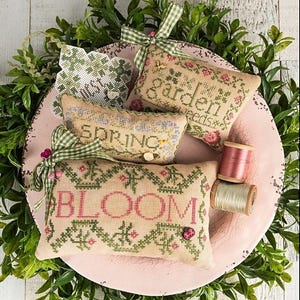 May include: A collection of embroidered spring-themed pillows with the words "SPRING," "BLOOM," and "GARDEN seeds" in pink and green. The pillows are arranged on a pink plate with greenery and spools of thread. The text "SPRING CARTOUCHES" is at the top.