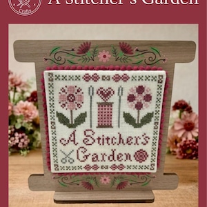 May include: A wooden cross-stitch display featuring a stitched design with flowers, a spool of thread, and the text "A Stitcher's Garden." The display has a decorative floral border and is set against a soft background.