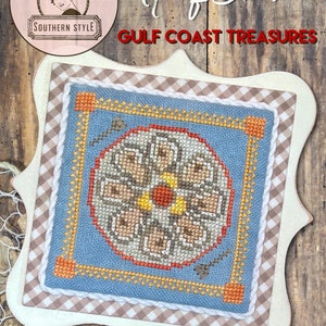 May include: A cross-stitch pattern of a half shell with a blue background and orange, yellow, and brown accents. The pattern is framed in a white frame with a brown and white checkered border. The text "Half Shell Gulf Coast Treasures" is written above the frame.