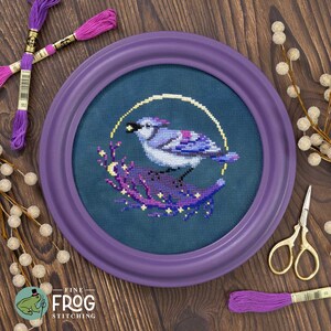 May include: A framed cross-stitch of a blue jay perched on a branch, surrounded by a gold halo and stars. The artwork is in a round purple frame. Embroidery floss and scissors are visible on a wooden surface. The Fine Frog Stitching logo is in the lower left.