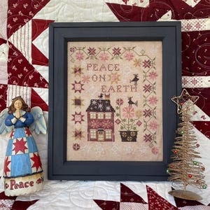 May include: A framed cross-stitch sampler with a winter scene. The sampler features a house with a red roof, a potted plant, and two birds. The text "Peace on Earth" is stitched in the center of the sampler.