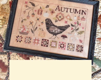 May include: A framed cross-stitch embroidery on tan fabric featuring a central black bird, the word AUTUMN, and various seasonal motifs including pumpkins, flowers, mushrooms, and geometric patterns in muted earth tones of brown, orange, and green.