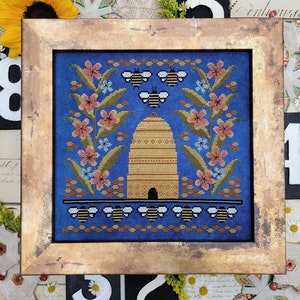 May include: A framed cross-stitch embroidery depicting a beehive with bees flying around it. The beehive is surrounded by flowers in shades of pink, blue, and green. The background is a deep blue color.