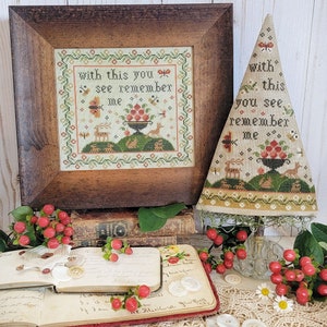 May include: A cross-stitch sampler with a brown wooden frame and a beige background. The sampler features a green hill with a basket of red apples, two deer, and a butterfly. The text on the sampler reads "with this you see remember me".