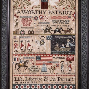 May include: A framed cross-stitch sampler with a patriotic theme. The design features historical figures, flags, and text including "A Worthy Patriot" and "Life, Liberty, & the Pursuit of Happiness." The color palette includes red, white, blue, and tan.