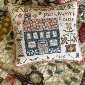May include: A square pincushion featuring a cross-stitch design of a house with the text "patchwork house" in brown thread. The house is navy blue with a patterned roof, surrounded by floral motifs and small sewing tools, finished with a pink trim.