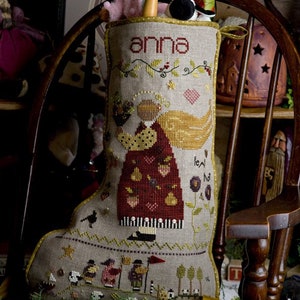 May include: A beige and brown Christmas stocking with an embroidered angel holding a basket of fruit. The stocking is personalized with the name "anna" and has the text "feed the hungry" embroidered on it.