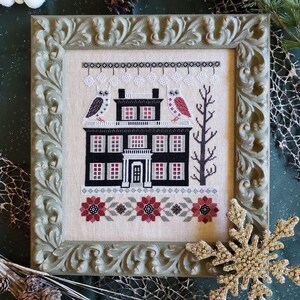 May include: A cross-stitch embroidery of a black and white house with red accents, two owls perched on the roof, and a snowy winter scene. The embroidery is framed in a green ornate frame.