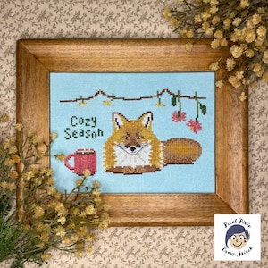 May include: Cross stitch pattern featuring a fox with a mug of hot cocoa and the text "Cozy Season". The pattern is framed in a wooden frame and surrounded by small yellow flowers.