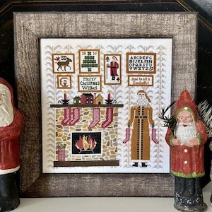 May include: A framed cross-stitch sampler with a Christmas theme. The sampler features a fireplace with stockings, a Santa figure, and the text "Merry Christmas Wishes" and "And to all a Goodnight". Two Santa figurines flank the framed sampler.