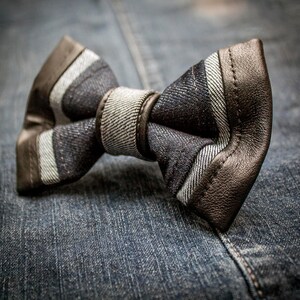 Może przedstawiać: Brązowy skórzany krawat muszkowy w denimowy wzór w białe paski. Muszka leży na niebieskim denimowym materiale.