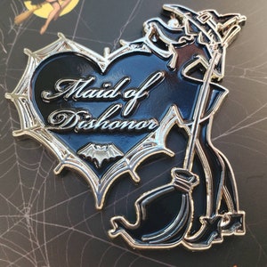 May include: A black and silver enamel pin featuring a witch holding a broom and a heart with the text "Maid of Dishonor" inside. The heart is surrounded by a silver spiderweb design.