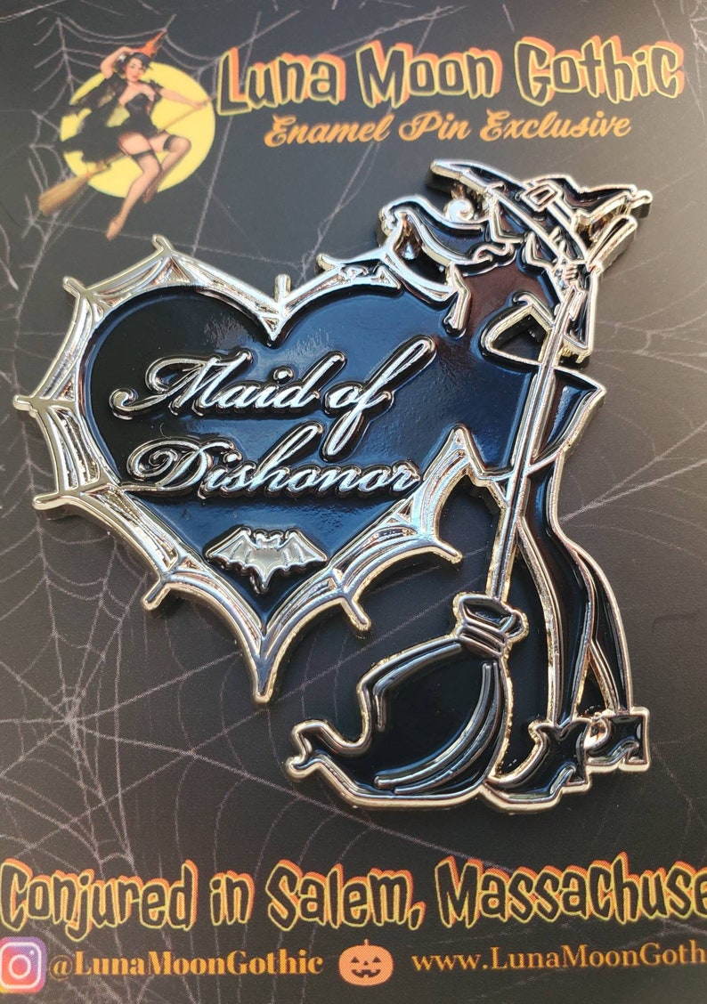 May include: A black and silver enamel pin featuring a witch holding a broom and a heart with the text "Maid of Dishonor" inside. The heart is surrounded by a silver spiderweb design.