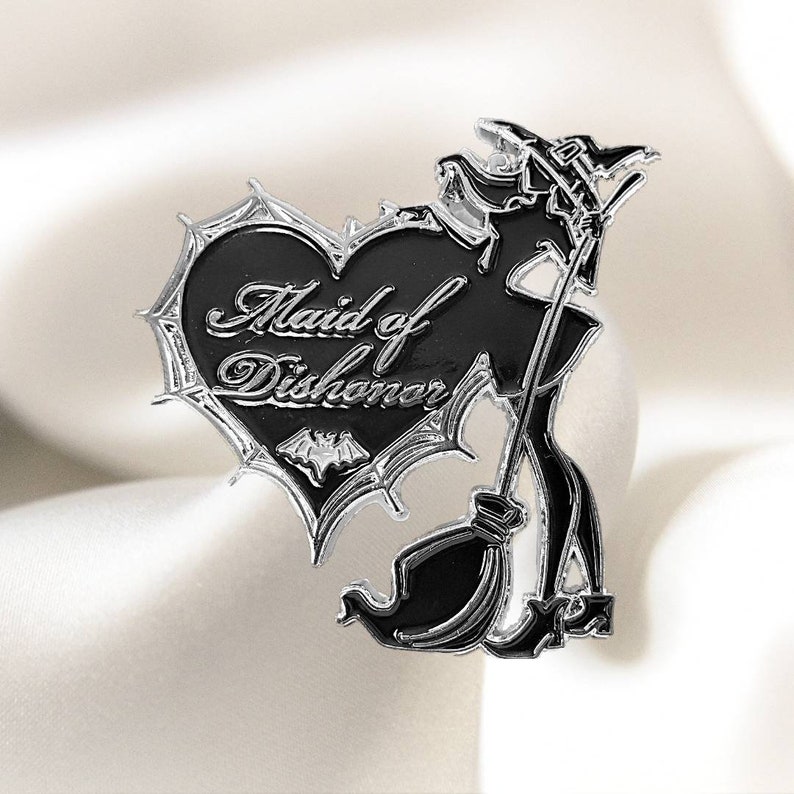 May include: Black and silver enamel pin featuring a witch with a broom and a heart with the text "Maid of Dishonor" in a gothic font.