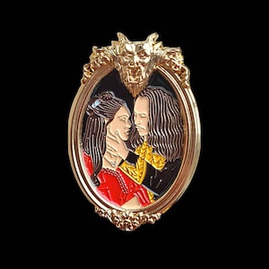 Könnte beinhalten: Goldfarbene Emaille-Anstecknadel mit einem Paar in romantischer Umarmung, gerahmt von einem dekorativen goldenen Rahmen mit einem Gargoyle-Design. Die Frau trägt ein rotes Kleid und der Mann trägt ein schwarzes und goldenes Outfit.