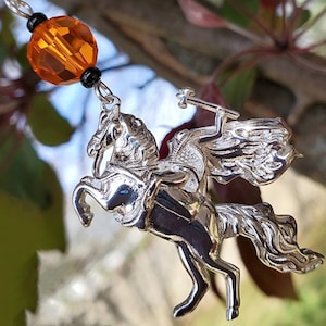 May include: Silver charm of a knight riding a horse, with a cape and sword. The charm is attached to a chain with an orange glass bead.