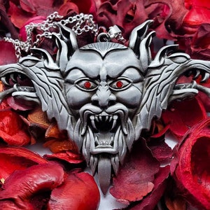 Może przedstawiać: Srebrny naszyjnik z zawieszką przedstawiającą szczegółowo zaprojektowaną, trójgłową wilczą głowę z czerwonymi oczami. Zawieszka ma kształt trójkąta i ma fakturowaną, prawie futrzastą powierzchnię. Naszyjnik jest na srebrnym łańcuszku.