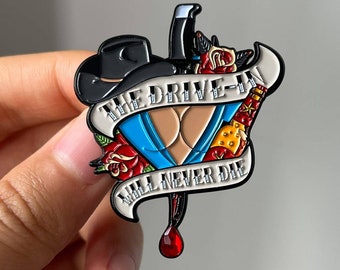 May include: Enamel pin featuring a black cowboy hat, a blue bikini top, and a dagger with a red gem. The pin includes the text "THE DRIVE-IN WILL NEVER DIE" on a banner, with red roses and a bottle.