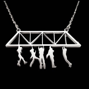 May include: A silver-colored necklace featuring a bridge design with five hanging figures. The bridge has a geometric truss structure. The figures are in various poses, suggesting movement. The necklace hangs on a silver chain against a black background.