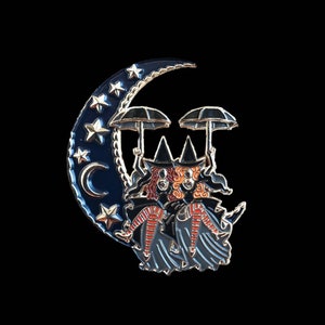 May include: Enamel pin depicting two witches with red hair and black hats, holding umbrellas, riding a crescent moon. The moon is dark blue with gold stars and a gold trim. The background is black.