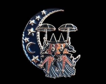 May include: Enamel pin featuring two witches with red hair and black hats, holding umbrellas, riding a crescent moon. The moon is dark blue with gold stars and a gold trim. The background is black.