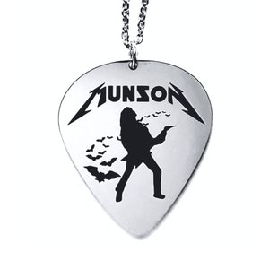 May include: A silver guitar pick necklace with a black silhouette of a person playing a guitar and bats flying around them. The pick has the word "MUNSON" written on it in a stylized font.