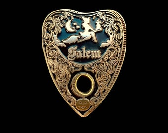 May include: A decorative planchette with a gold-colored frame and a black center. The planchette features a witch on a broom, a crescent moon, and the word "Salem". A pumpkin is at the bottom.