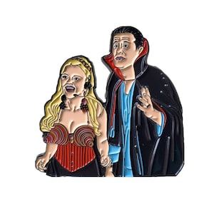 May include: A black and red enamel pin featuring a cartoon couple dressed in a vampire costume and a red and white striped corset. The man has a black cape and fangs. The woman has blonde hair and a microphone.