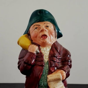 Puede incluir: Figura de cerámica de un hombre con vestimenta histórica. Lleva un sombrero verde oscuro, un abrigo burdeos, un chaleco verde y pantalones amarillos. La figura sostiene un objeto amarillo y un pergamino.