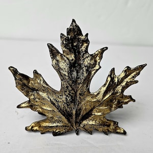 Könnte beinhalten: Ein dekoratives Metallblatt im Ahornblatt-Design. Das Blatt ist überwiegend goldfarben mit schwarzen Akzenten und weist komplizierte Adern und eine strukturierte Oberfläche auf. Kann als dekoratives Element oder als Schrankgriff verwendet werden.