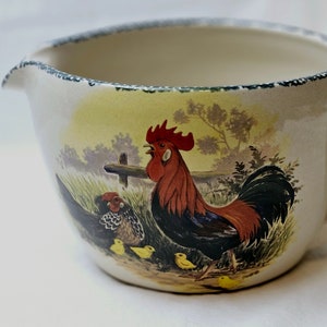 以下が含まれることがあります： 鶏とひよこのデザインが施されたセラミックピッチャー。ピッチャーは白地に黄色、緑色、茶色のデザインが施されています。鶏は赤色、黒色、黄色です。ひよこは黄色です。