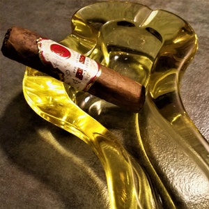 May include: A yellow glass ashtray with a cigar resting inside. The cigar has a white band with red and gold accents and the text "El Sol" printed on it.