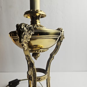 May include: A polished brass table lamp featuring a central fluted column and a bowl-shaped body adorned with three sculpted ram head motifs. The lamp stands on a circular base with three curved legs and includes a black electrical cord and switch.