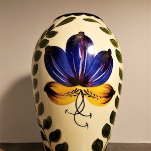 Może przedstawiać: Kremowy ceramiczny wazon z dużym niebiesko-żółtym kwiatowym wzorem. Wazon jest ozdobiony zielonymi wzorami liści i czarnymi akcentami. Wazon ma zaokrąglony kształt i prawdopodobnie służy do celów dekoracyjnych. Ma około 30 cm wysokości.