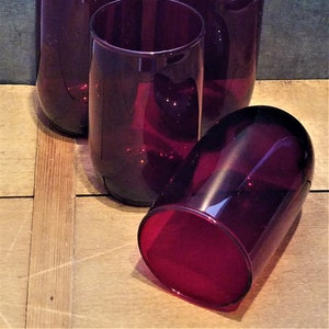 May include: Four clear, ruby red glass tumblers. The tumblers are stacked on a wooden surface.