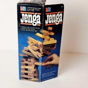 Könnte beinhalten: Eine Vintage-Jenga-Spielbox mit dem Spielnamen in Weiß auf einem schwarzen und blauen Hintergrund. Die Box zeigt einen Turm aus Holzblöcken und eine Hand, die einen Block entfernt. Das Spiel ist für Kinder ab 6 Jahren geeignet.