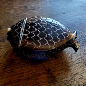 May include: A decorative fish-shaped box with a hinged lid. The box is dark brown with a scale pattern and a gold-colored trim. The fish has a small, ivory-colored accent near the mouth and a silver-colored clasp.