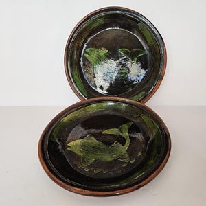 May include: Two handmade ceramic bowls. The bowls are dark brown with a green fish design in the center. The rims are a lighter brown. The fish are green. The bowls are round and have a rustic appearance.